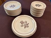 Set of 12 tan stoneware dessert plates with brown floral stencil design by Pfaltzgraff Village. Plates stacked in two groups showing front and side views.