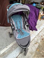 Front angle view of single baby stroller with canopy, padded teal and gray seat, and front swivel wheel on concrete floor.