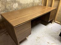 Photo of large wooden office desk with drawers on both sides, sitting on rollers. Right leg shows damage. Desk located in basement with concrete walls and floor.
