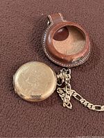 Close-up of the closed gold-tone Elgin pocket watch with engraved outer cover and metal chain attached to a brown leather circular-front case.