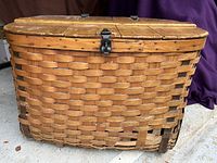 Front view of large rectangular wooden woven basket with hinged lid and metal latch, showing woven wood slats with metal reinforcement and some wear at base.