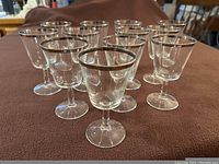 Twelve clear glasses with silver rims on a brown surface, arranged in loose group.