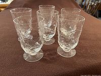 Six medium-sized clear wavy textured glass drinking glasses, each with short stems and round bases, arranged on a brown fabric surface.