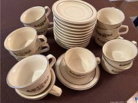 Image showing 12 tan coffee cups with brown stencil design stacked and grouped around 12 matching saucers on a flat surface.
