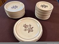Two stacks of Pfaltzgraff Village Stoneware plates, one stack of larger dinner plates and one stack of smaller plates, showing brown floral stencil design on tan background, some visible chips on edges.