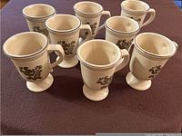 Eight tan Pfaltzgraff Village Stoneware mugs with brown stencil floral designs on a dark cloth.