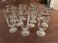 Photo shows eleven clear parfait glasses arranged in rows on a brown surface. Each glass has a silver rim and a stem with a circular base.