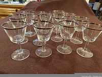 Twelve small clear glass drinking glasses with silver rims arranged on a brown cloth surface, showing their general size and design.