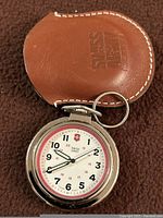 Photo showing mens Swiss Army pocket watch model 24721 with clear view of white dial, black and red numerals, and silver-tone case resting on brown surface with brown leather pouch behind it embossed with Swiss Army.