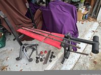 Weight bench with red padded segments on black frame, with various black metal poles and anchors on the floor beside it.