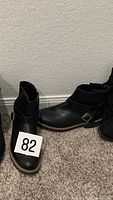 Front view of pair of black leather ankle boots with cross straps and buckle, showing toe, ankle opening, and sole color contrast.