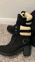 Side view of a black lace-up boot with cream wool ankle lining, buckle strap on the side, and stacked chunky heel resting on carpet.