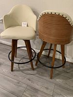 Two new bar stools shown side by side on a wood floor. One stool is oriented facing forward and the other shows the back side, highlighting the bamboo wooden styled curved backrest with horizontal lines and studded trim. Both have padded cream leather seats with wooden legs and a black metal circular footrest.