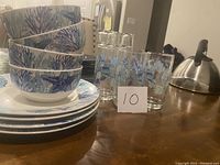 Photo showing four stacked ceramic bowls and four stacked ceramic plates with blue coral design, next to four clear glasses with blue sea life motif, on a wooden table.