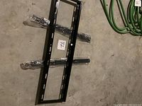 Black metal TV wall mount bracket laid on a concrete floor with green cables nearby. The item has multiple holes for mounting and wrapped parts in plastic.