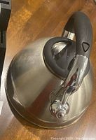 Photo showing stainless steel whistle kettle with curved black handle and chrome spout cover on wooden surface.
