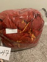 King size comforter and bed skirt folded and packed inside a clear plastic bag. The comforter is red with yellow and brown floral or leaf designs.