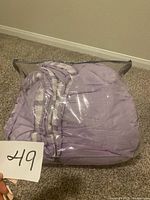 Packaged king size purple comforter inside a clear plastic bag on a carpeted floor, held with a lot number card marked 49.
