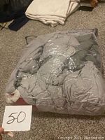 Grey comforter wrapped in clear plastic packaging lying on carpeted floor with a tag showing number 50.