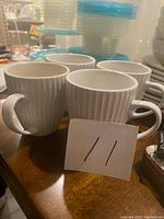 Four white ribbed coffee mugs displayed on a wooden table, showing front and side views with visible ribbing and handles.