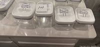 Five clear square pantry storage containers with white lids and clear plastic handle closures placed on a countertop.