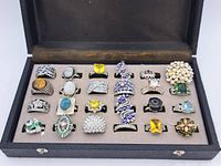 Overview of the 24 assorted gemstone rings in a display case including tiger's eye, moonstone, pearl cluster, amethyst, citrine, and green stones.