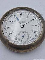 Close-up of watch face showing white dial, black Roman numerals, blue hour hand, black minute hand, and small seconds dial at 6 o'clock. Metal case with patina visible.