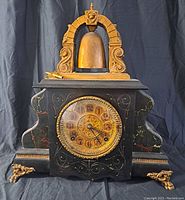 Front view of the antique Bell O Matic clock showing ornate bell on top, clock face with decorative gold-tone bezel and black and gold numbered dial, black case with gold trim and ornamental feet.