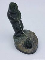 Front view of the jade fisherman figure holding a metal wire fishing line, standing on a textured jade stone base with carved fish in a pond depression.
