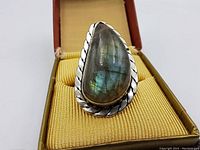 Front view of the pear-shaped labradorite ring set in a twisted rope detailed sterling silver bezel placed in an open jewelry box with a yellow fabric interior.