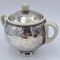 Front and side view of the round porcelain container wrapped in hammered sterling silver with glazed porcelain handles and knob on lid.