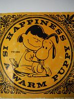 Close-up photo of the cover of 'Happiness Is A Warm Puppy,' featuring Lucy hugging Snoopy on a yellow-orange background with stylized text and corner decorations.