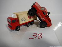 Photo of red Coca-Cola delivery box truck with cream cargo box and logos, posed with red pickup truck on top