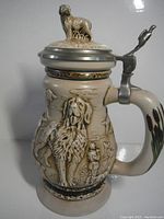 Front and side view of ceramic hunting dog beer stein with relief and pewter lid showing dog figurine on top.