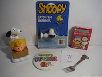 Full lot showing Snoopy bobber figurine with Sally in a basket, unopened Peanuts playing cards, 1969 Peanuts popables button, and Snoopy fork.
