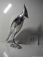 Front and side views of a large polished metal cardinal sculpture with smooth surface and stylized features, posed on a flat base with a number 67 card nearby.
