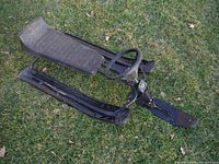 Side angle photo of the black GT SnowRacer sled on grass, showing the steering wheel, padded seat, and three metal runners.