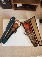 Photo showing an antique ukulele in a teal-lined case and a vintage violin with bow in a beige-lined case, both on a white surface.