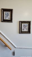 Two framed art prints hung on a wall above a staircase railing shows size and placement of the prints
