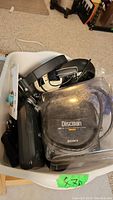Photo showing a plastic bin holding a Sony Discman portable CD player, black over-ear headphones, and other black audio devices.