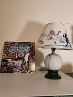 Full view of vintage baseball-themed lamp with cartoon baseball player illustrations on the shade and baseball base, with the baseball-themed LP visible behind it showing baseball memorabilia.