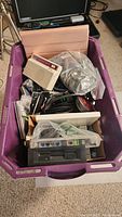 Overhead image showing a purple plastic bin filled with various computer accessories including cables, adapters, an external device, a mouse, floppy disk drives, and other peripherals.