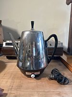 Front view of vintage stainless steel electric kettle with black handle and coiled cord resting beside it on wooden surface.