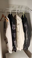 Four men's jackets and sweaters hanging on a clothing rack inside a closet, various colors and materials.