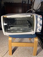 Front view of Black & Decker toaster oven placed on a small wooden stool with blue mat, visible interior rack and crumb tray, three control knobs on right, some dirt on door glass.