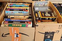 Two cardboard boxes filled with approximately 28 VHS cassettes featuring several Disney movie titles and other movies.
