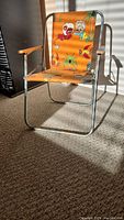 Full view of vintage children's picnic chair with orange cartoon fabric and metal frame with wooden armrests