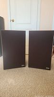Front view of two black grill cloth covered SOTA Seakers speakers standing side by side on beige carpet.