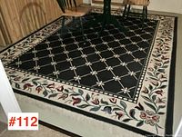 Full view of the rectangular carpet with black background and floral border with partial view of a green leg dining set on carpet.