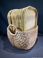 15 rectangular bamboo trays with woven bamboo surfaces stacked inside a natural woven rattan basket with side handles.
